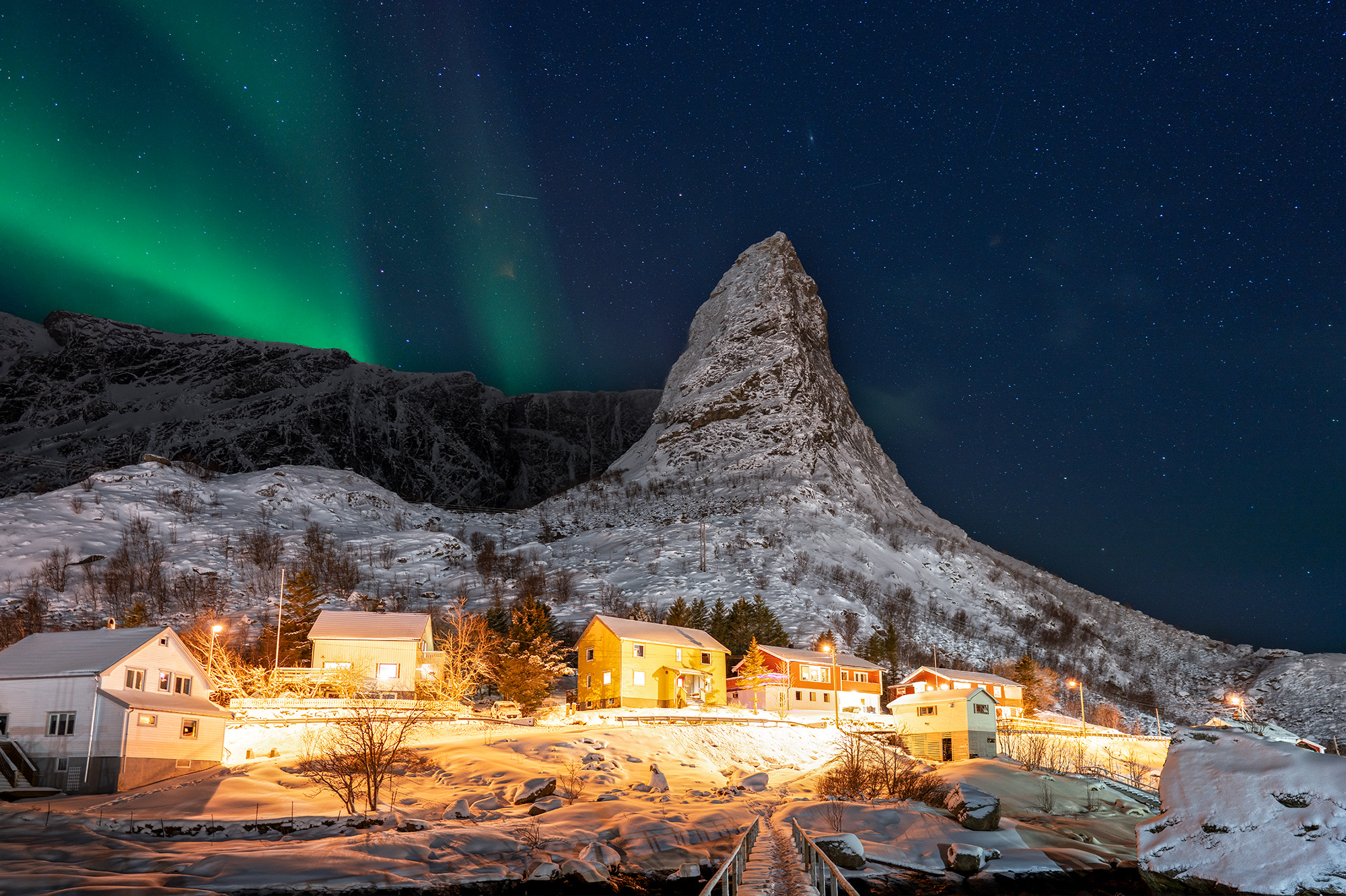 Northern Lights Over Reine’s Winter Night This image captures a serene winter night in Reine, Norway, where warmly lit homes contrast against the cold, snowy landscape...