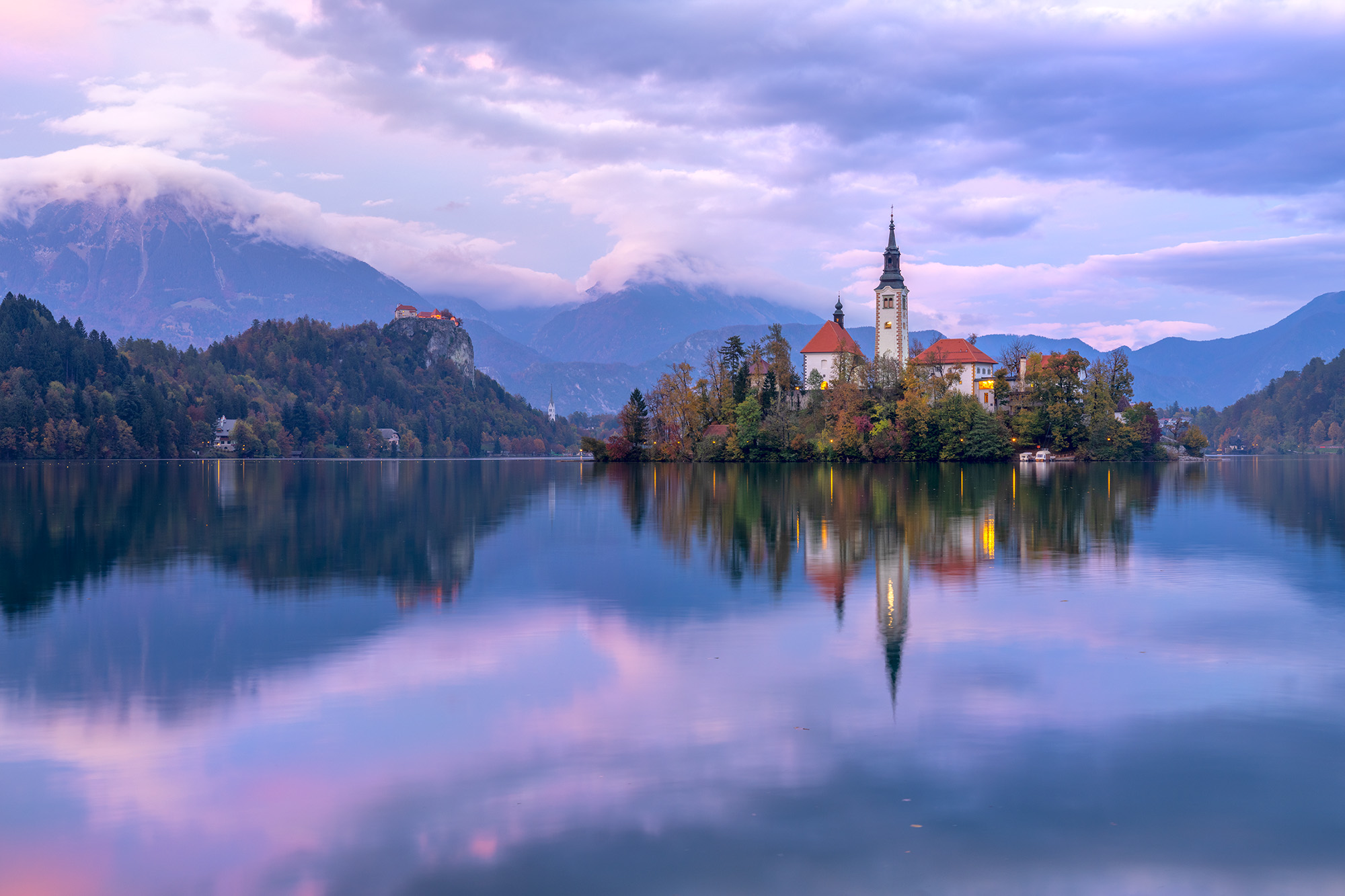 As daylight fades on Lake Bled in Slovenia, the Pilgrimage Church gracefully mirrors upon the tranquil waters. In the distance...
