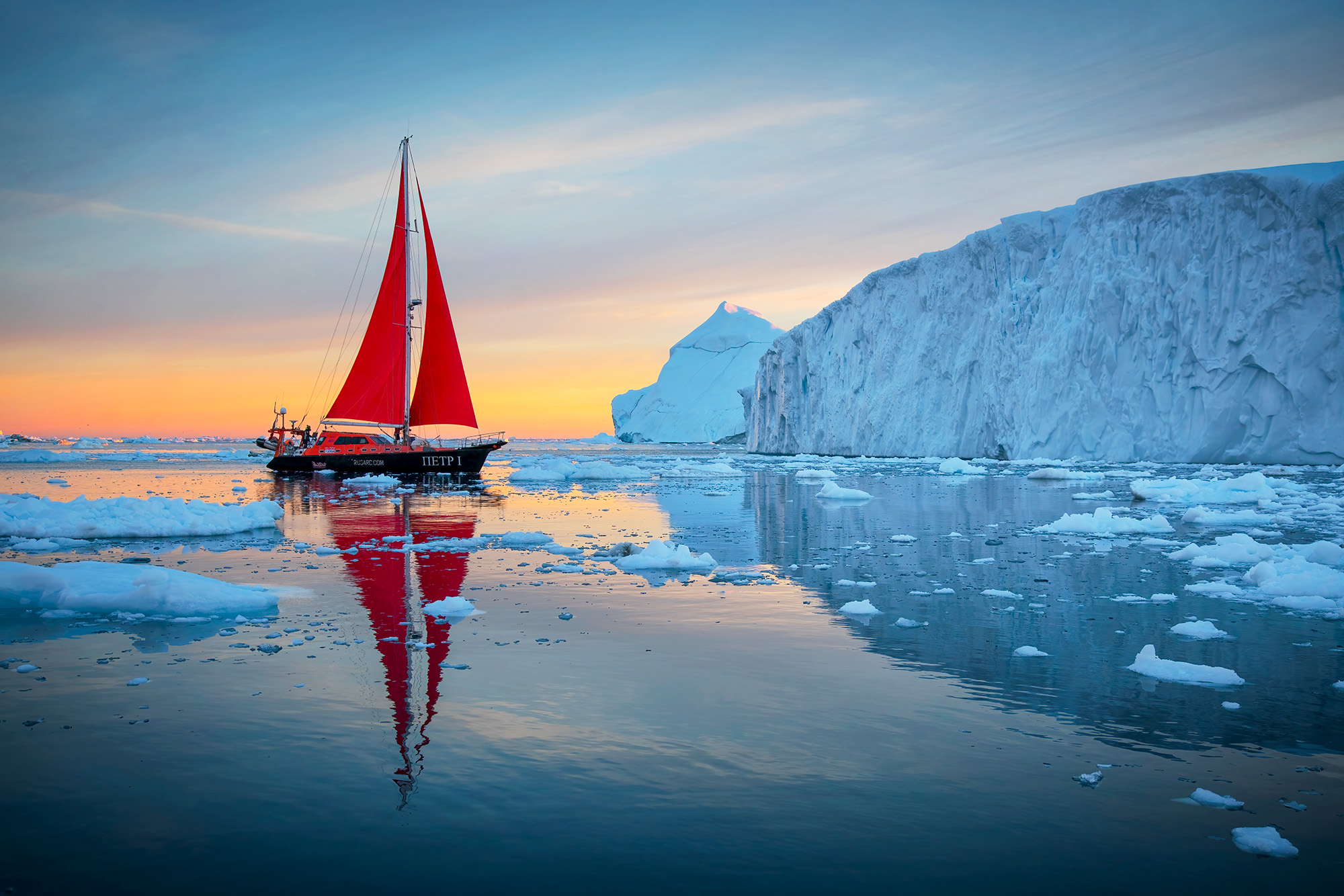 Captured in Disko Bay near Ilulissat, Greenland, this image portrays a sailboat with vibrant red sails gracefully approaching...