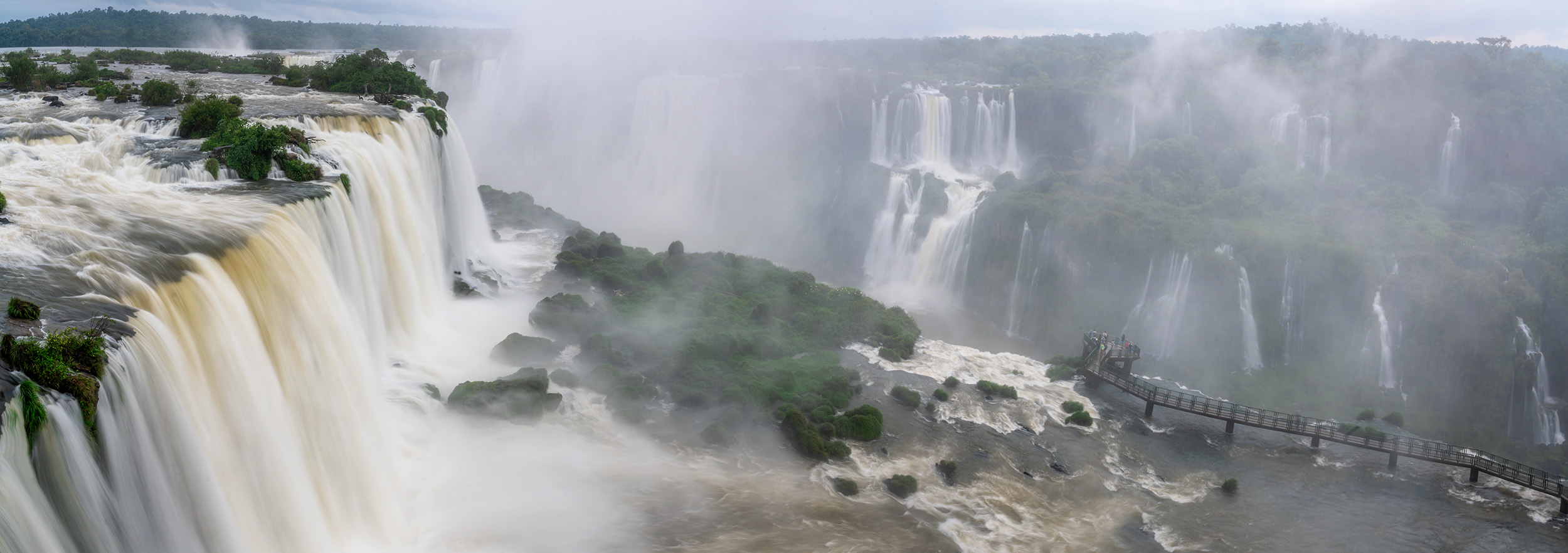 Veiled Power Panoramic In the heart of Iguazu Falls, Brazil, "Veiled Power" captures the majestic falls under moody skies and the shroud of an approaching...