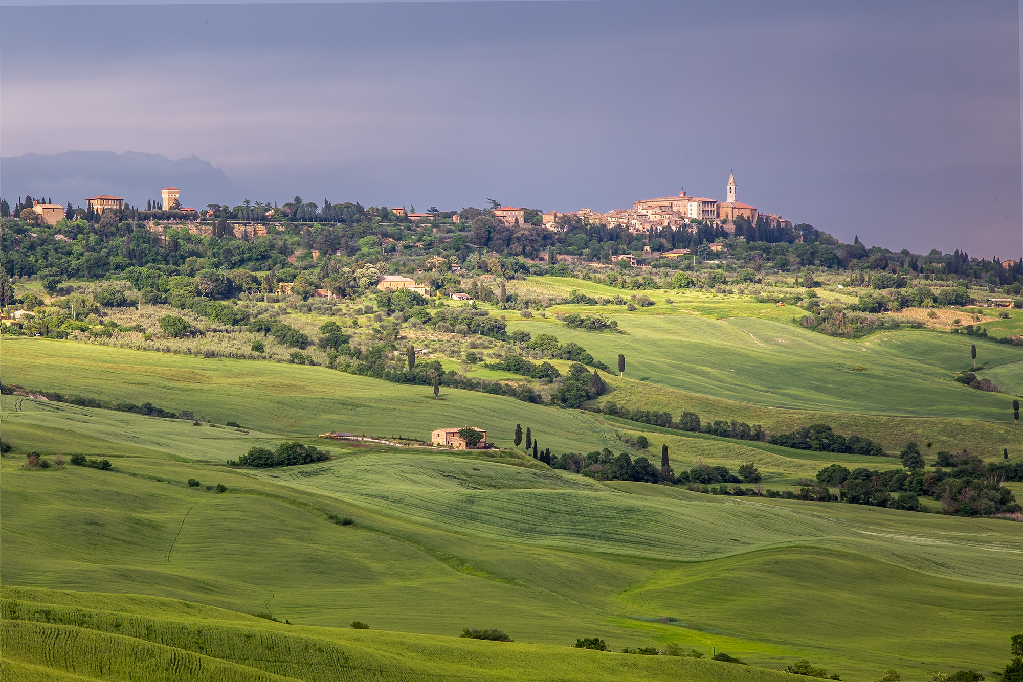 In this image, the drama of a Tuscan thunderstorm unfolds. Vast grasslands stretch towards the distant town of Pienza, bathed...