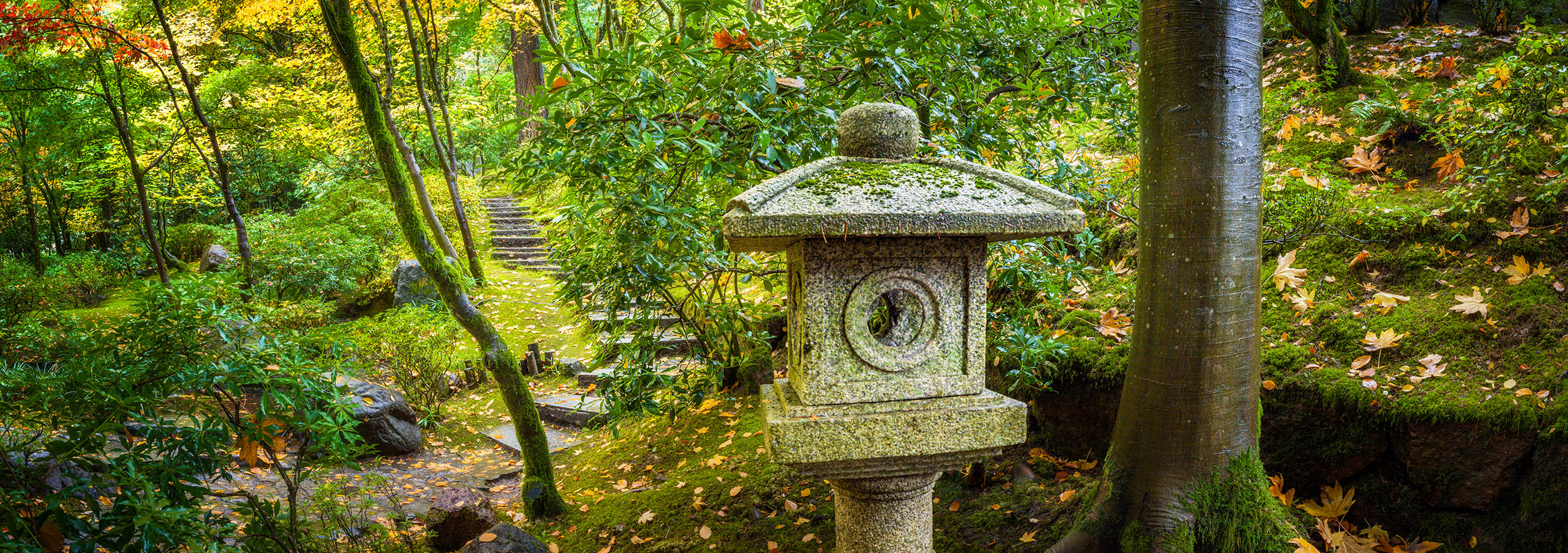This image, a panoramic composition from the Portland Japanese Garden in Oregon, showcases a stone lantern and a set of stone...