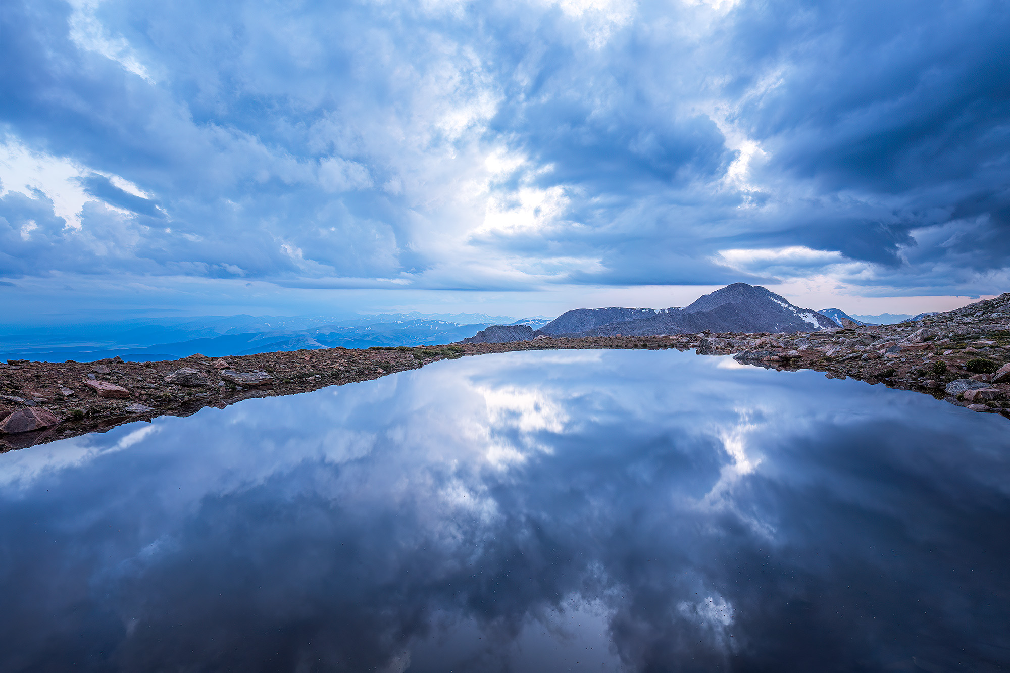 This image captures the an alpine escape atop Mount Evans, Colorado. A serene tarn rests amidst the rugged landscape, mirroring...