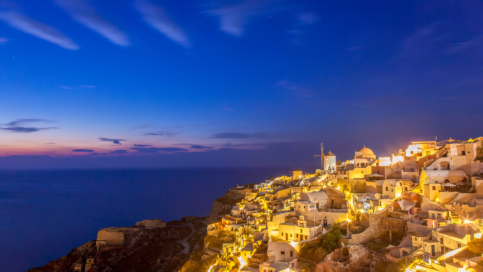 Captured during the late blue hour, this image offers a sweeping panorama of Oia, Santorini, Greece. On the left, the charming...