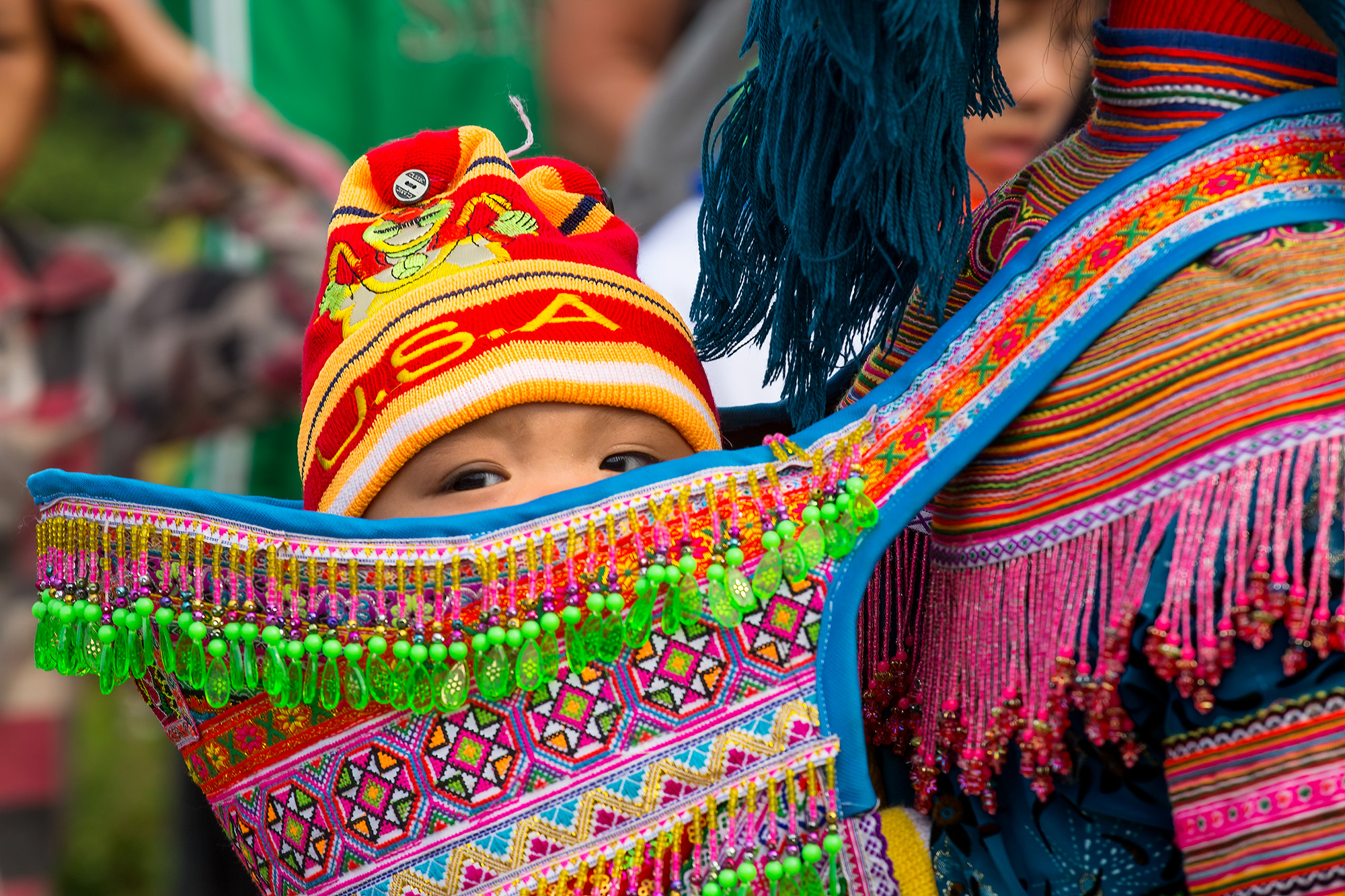 Innocence Adorned Amidst the vibrant tapestry of Bac Ha, Lao Cai, Vietnam, this image centers on a child adorned in a brilliantly decorated Hmong...