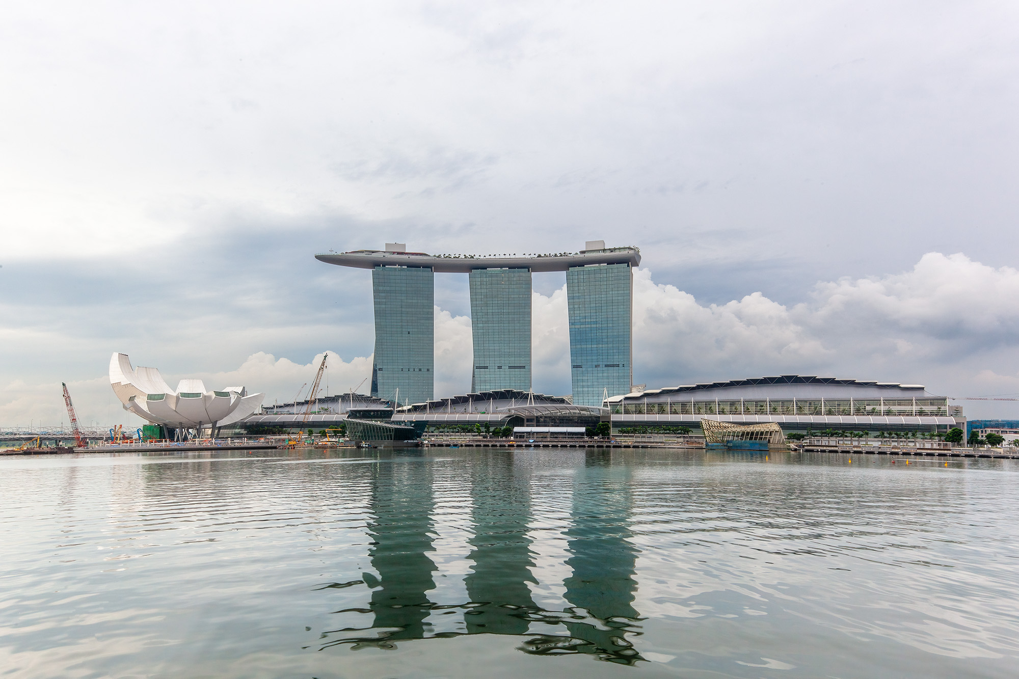 This image captures the iconic Marina Bay Sands Hotel and Casino on a cloudy day, as viewed from the harbor. The reflection of...