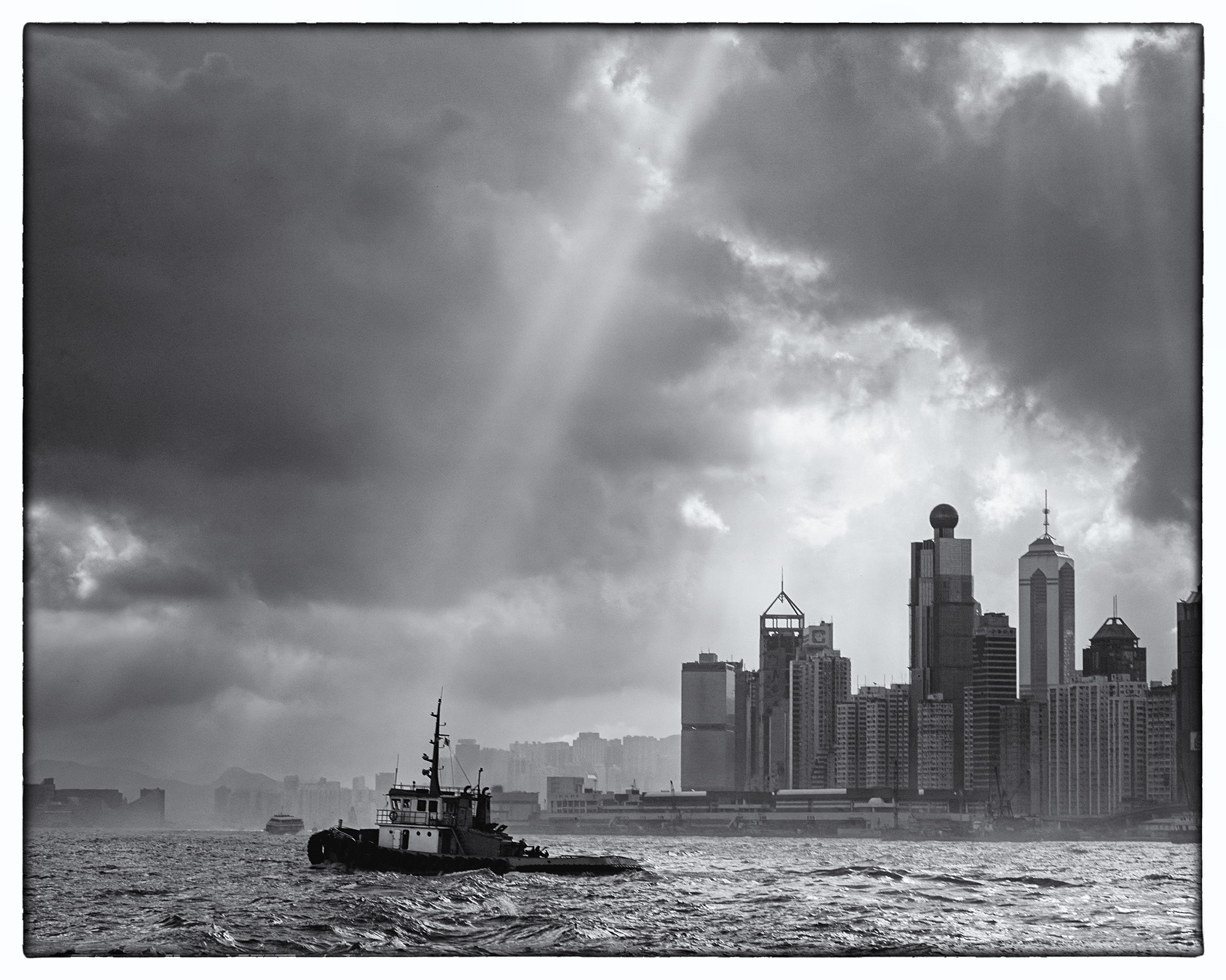 This striking black and white image captures the essence of bustling Hong Kong Harbor. A sturdy tugboat plies the waters, guided...